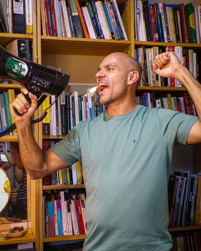 Fernando Viana grita num megafone e ergue o punho, diante de uma estante cheia de livros.